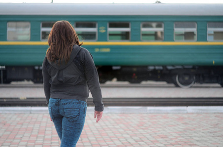 一个年轻的红头发女孩站在铁路站台上，看着离去的火车。 那个女人赶火车迟到了。 背面视图
