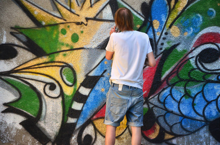 在旧混凝土墙上画涂鸦图案的过程中的照片。 年轻的长发金发男人画了一幅不同颜色的抽象画。 街头艺术和破坏概念