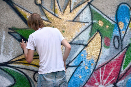 在旧混凝土墙上画涂鸦图案的过程中的照片。 年轻的长发金发男人画了一幅不同颜色的抽象画。 街头艺术和破坏概念