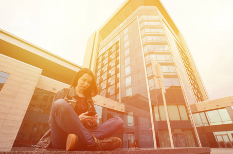 一个穿牛仔裤衣服的年轻女孩，用智能手机坐在蓝色办公楼的花岗岩栏杆上。 使用现代技术互联网和接收在线信息的概念