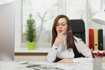 美丽的严重和全神贯注的棕色头发的业务妇女在西装和眼镜坐在办公桌与片剂, 工作在计算机与现代显示器与文件在轻型办公室, 看一边