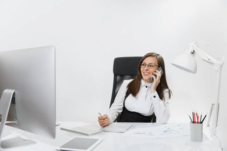 漂亮的微笑商务妇女坐在办公桌前, 在现代计算机与文件在轻型办公室, 在手机上交谈, 进行愉快的谈话在白色背景