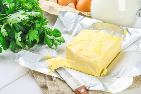 一条黄油在木板上切成块, 上面有一把小刀, 周围是牛奶鸡蛋和欧芹。烹调配料