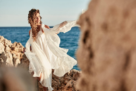 一幅美丽的年轻模特的肖像，留着长长的黑发，穿着长长的白色连衣裙，在夏天的深蓝色海洋附近的岩石上摆着姿势。