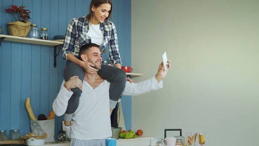 快乐的年轻夫妇有乐趣和聊天在线视频电话在家里的厨房。女孩坐在男朋友脖子上, 而他手持智能手机