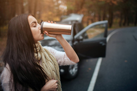 在车旁拿着一次性杯咖啡的女人。关闭手。秋天的概念。秋天森林旅途由汽车