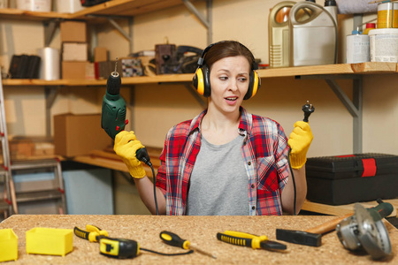 困惑的年轻女子在格子衬衫灰色 t恤噪音绝缘耳机黄色手套工作在木工车间的木制桌位, 不同的工具, 电钻, 电线