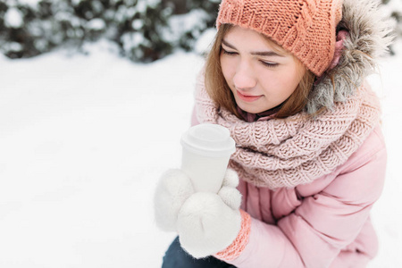 一个美丽的年轻女孩的肖像在白色针织手套, 户外, 拿着一个热饮料的纸杯, 明亮的冬日. 女人微笑着, 是幸福的, 关闭, 在第一