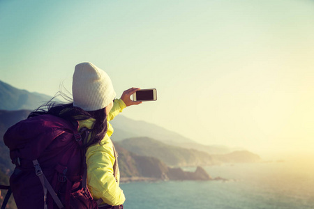 年轻女性徒步旅行者在海边拍照时使用智能手机