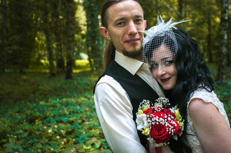 黑发新娘妻子和她未婚夫在一个绿色的秋天公园散步。 她穿着一件带有花束的复古长裙，穿着刺的脸藏在面纱下