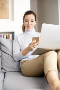 自信的成熟女人在她的手机上阅读信息，并在家里使用笔记本电脑。