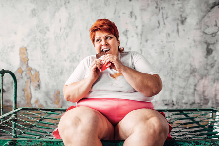 超重的女人坐在生锈的床上，吃巧克力懒惰和肥胖的概念。 不健康的食物