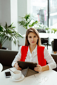 严格的商务女士坐在咖啡馆的一张小桌子上，在平板电脑上使用互联网，喝咖啡。 在商务中心午餐时穿着白色衬衫和红色夹克的女人。