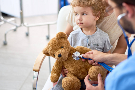 医生办公室里可爱的卷发孩子的肖像，拿着泰迪熊玩具，儿科医生用听诊器听心跳