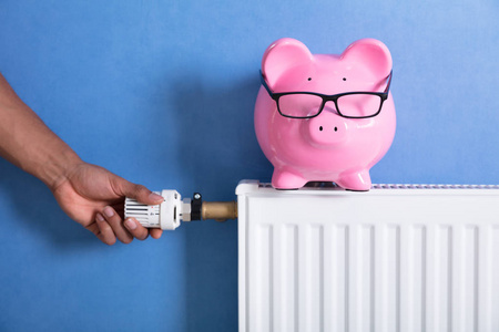 s Hand Adjusting Thermostat With Piggy Bank On Radiator