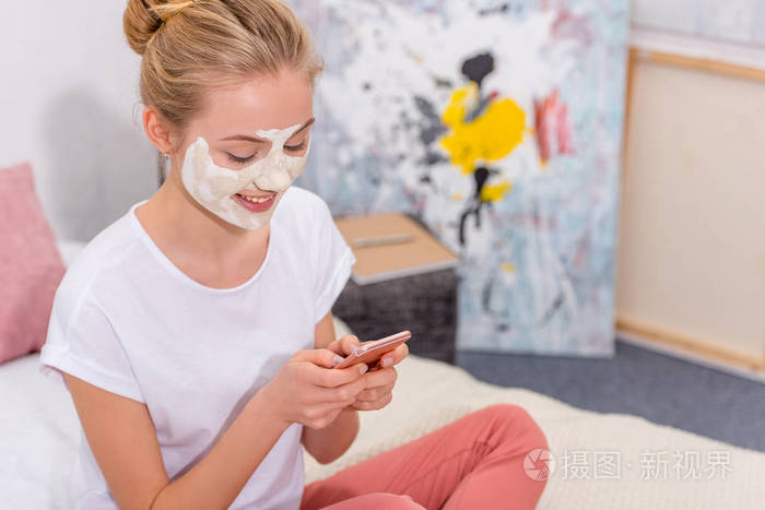 幸福的年轻女人,带着白色粘土面膜,在家坐在床上,用智能手机做面膜