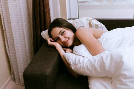 可爱的微笑的女孩躺在床下一条白色的毯子, 不想起床