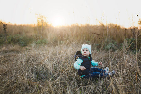 小可爱的孩子男孩在绿松石帽子, 背心, 牛仔裤坐在干燥的高草背景上春季或秋季自然。小孩儿子亲子家庭日5月15日爱父母的孩子概念