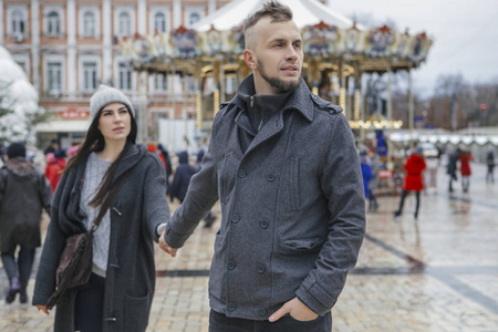 美丽美丽的高加索女人和一个男人在欧洲城市散步在冬天。 夫妻俩穿着休闲的灰色衣服，拥抱在一起。 天气寒冷，背景上有圣诞灯