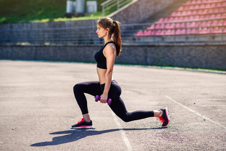 健身女孩与紫外线哑铃做热身之前，训练伸展身体肌肉。 女性运动员为心脏锻炼准备腿部。 活跃的女孩在户外做运动。