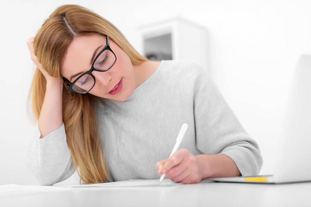 一个戴眼镜的金发女郎在办公桌旁的笔记本电脑旁边工作。年轻妇女在办公室或学习