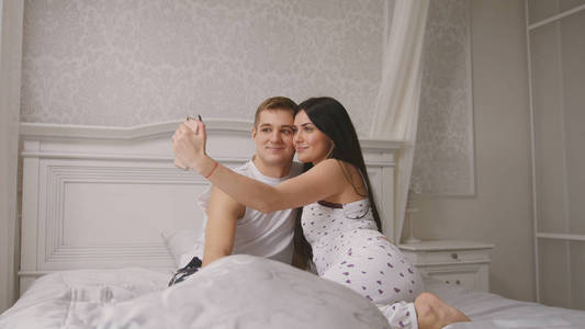 快乐欢快的情侣在床上做自拍, 年轻迷人的男人和女孩坐在睡衣