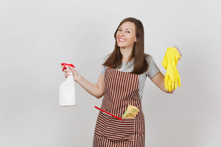年轻微笑的家庭主妇在条纹围裙与清洁抹布刮在白色背景的口袋隔离。女管家手持喷雾瓶, 用干净的液体黄色手套。瓶拷贝空间