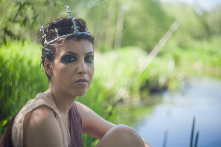一个棕色的裙子，头上戴着一顶皇冠，一个苗条的黑发女人坐在森林池塘的岸边