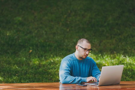 年轻成功的认真聪明的男子商人或学生在休闲蓝衬衫眼镜坐在桌子上的手机在城市公园使用便携式电脑在户外工作。移动办公的概念。复制空间