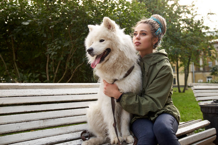 一个迷人的年轻女子与辫子坐在公园长椅上, 她雪白的萨摩耶狗。女主人亲切地拥抱她的宠物, 并按他给她