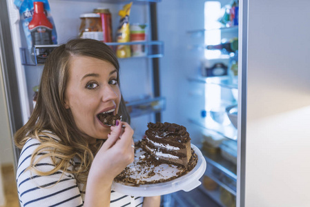 靠近站在开放冰箱前吃蛋糕的年轻女人。 冰箱前的女人手里拿着甜点盘上的蛋糕。 在冰箱前吃不健康巧克力蛋糕的女人