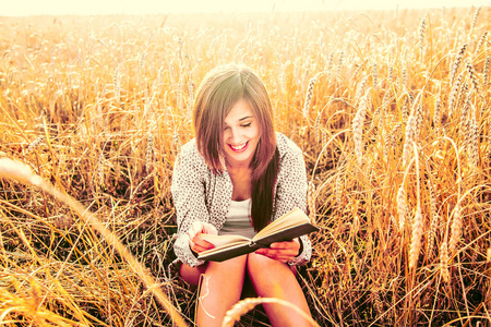 年轻女孩坐在金色收割小麦的肖像在黄色的夕阳田野。 女人读书