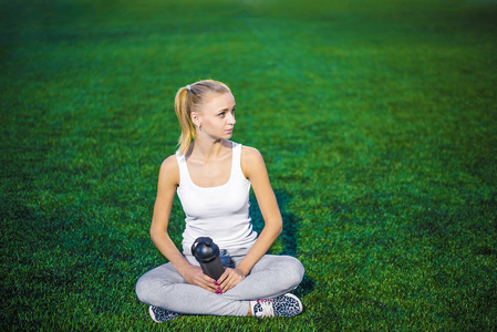 一个金发碧眼的女孩坐在户外的春绿草地上，拿着水瓶