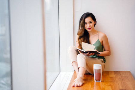 一位年轻的中国亚裔妇女早上坐在公寓的窗户旁，一边喝着健康的奶昔，一边读着一本书。 她即将开始她的一天，正在享受一个安静的时刻。