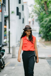 一位年轻的印度亚洲模特戴着一顶联邦制帽子站在亚洲的一条小巷里的肖像。 她在白天微笑，看着远方，优雅漂亮和高大。