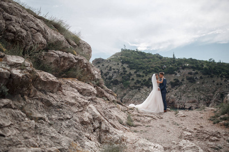 一对美丽的新娘和新郎在他们的婚礼上沿着海边的岩石海岸散步。年轻的夫妇在山里的春天，蜜月，快乐的微笑的新娘和新郎一起在山区度过时光