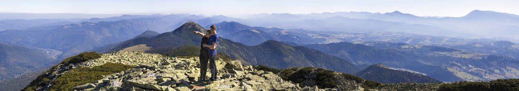 年轻夫妇徒步在大山。男人和女人站在山顶上看着下面美丽的风景。在温暖的夏季天气中高岩石丘陵的全景。体育和活动