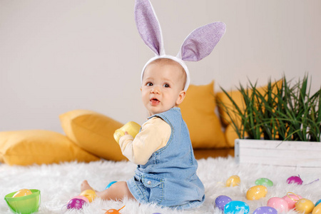 可爱的白种人女婴穿着牛仔裤，穿着紫色的兔子耳朵坐在工作室里。 孩子玩复活节彩蛋庆祝传统的基督教节日。