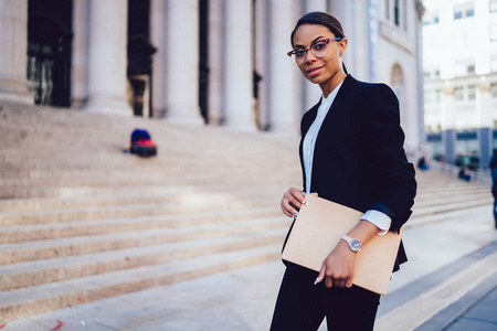 半长肖像的女非洲裔美国执行经理，穿着正式优雅的西装，看着相机，同时站在办公楼附近工作。 商业资料复制空间范围