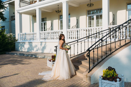 优雅的新娘与婚礼鲜花花束, 迷人的女人在婚纱礼服。穿着白色连衣裙的年轻女孩去了一所美丽的白宫的楼梯。幸福的新婚女人。婚礼化妆新娘