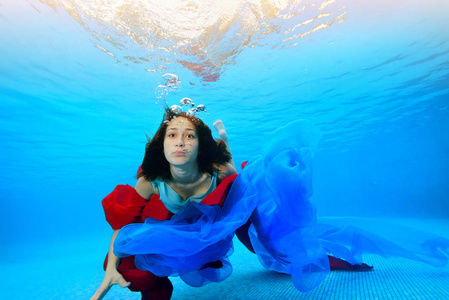 一个十几岁的女孩带着红色和蓝色的布游在水下，靠近底部，看着相机。 肖像。 在水下射击。 水平方向