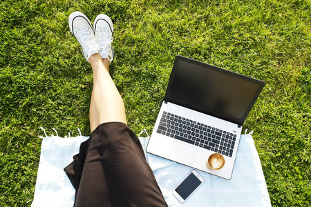 坐在绿色草坪上的大学生女孩在她的笔记本电脑上做作业。年轻女子坐在公园写博客, 写文章, eshopping, 社交网络在她的电脑