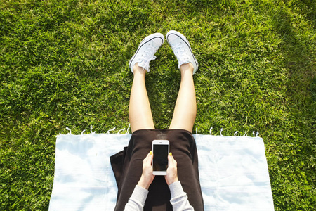 坐在绿色草坪上的大学生女孩在她的智能手机设备上发短信。年轻女子坐在公园写博客, 冲浪, eshopping, 社交网络, 手机小