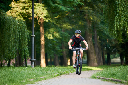 在夏季，骑自行车沿着公园小巷骑自行车时，穿着专业运动服和防护头盔的男自行车手看着一边。 实现目标