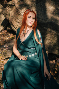 秋天的森林里走着一个美丽的红发女孩，她穿着一件绿松石的连衣裙，头上戴着一顶。在精灵的形象里。它在山上的大岩石上。太阳眩光。幻想