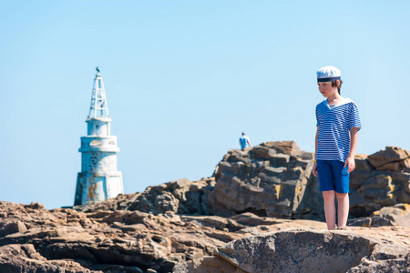 穿着条纹T恤和岩石上的帽子的男孩。 在背景上矗立着一座灯塔。