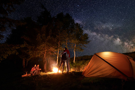 在橙色帐篷附近的营火周围露营的游客。一对拥抱在一起，第二对坐在星空下的原木上，银河坐在树的背景上