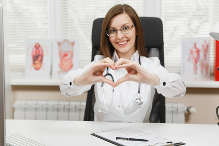 微笑的女医生坐在办公桌前, 用手显示形状的心脏, 与医疗文件在医院的轻型办公室工作。妇女在咨询室的医疗礼服。保健医学概念