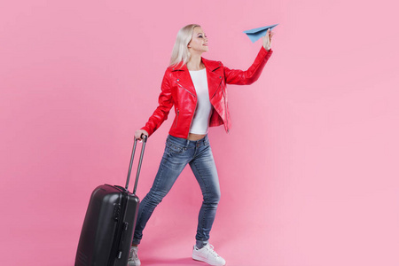 愉快的年轻妇女与旅行手提箱发射纸飞机。粉红色背景金发女郎, 概念