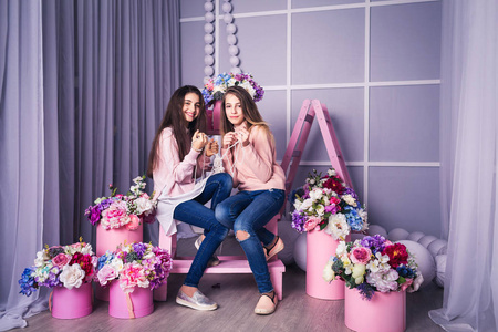 两个穿着牛仔裤和粉红色毛衣的漂亮女孩在画室里拿着珠子装饰着篮子里的花。